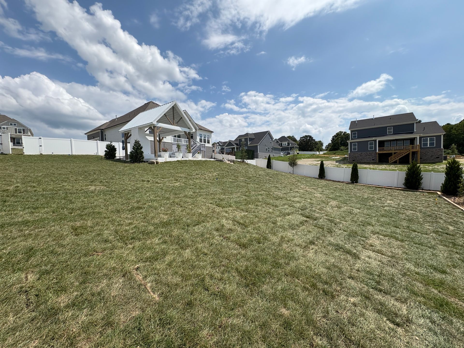 A wide, grassy lawn with a white fence, and houses under a partly cloudy blue sky.