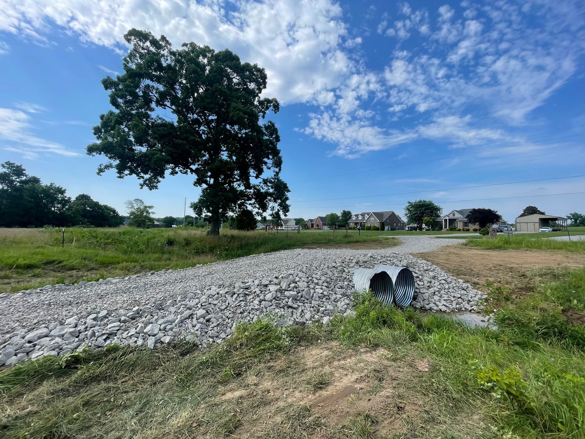 A rocky drainage area with two pipes, a tree, grassy field, and a blue sky with clouds. Houses in the distance.