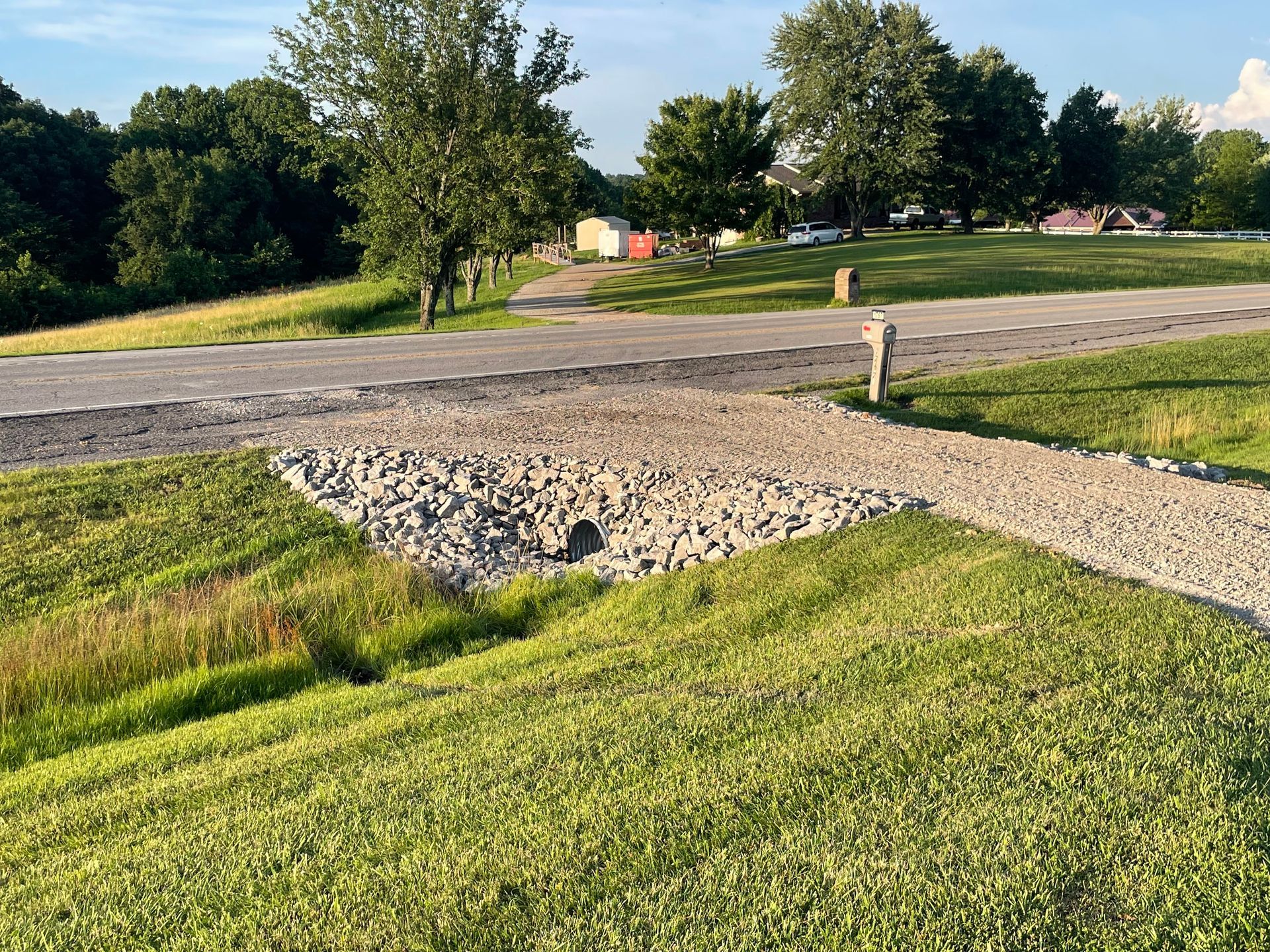 A grassy area with a gravel road, culvert, and trees in the background.