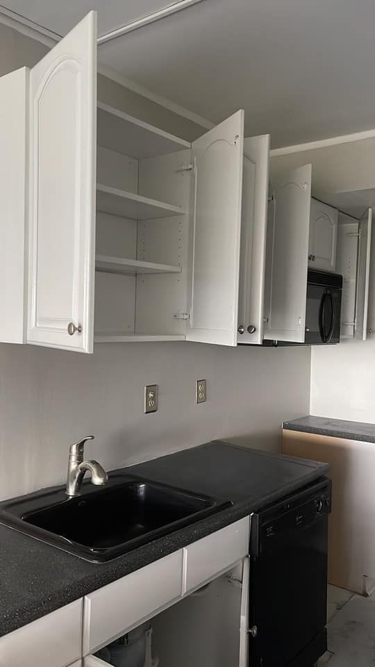 A kitchen counter with a black sink and dishwasher, featuring white cabinets with open doors against a light wall.