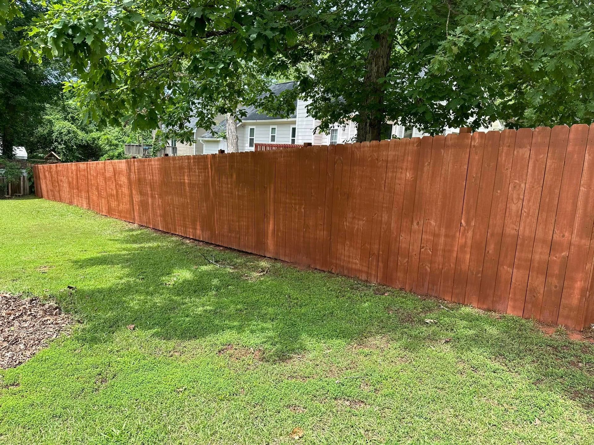A tall, stained brown wooden privacy fence borders a lush green lawn on a sunny day.