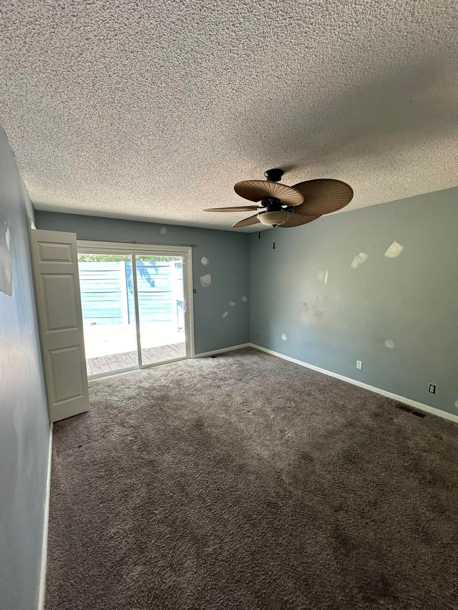 An empty bedroom with gray walls, a ceiling fan, brown carpet, and a sliding glass door leading to a patio.
