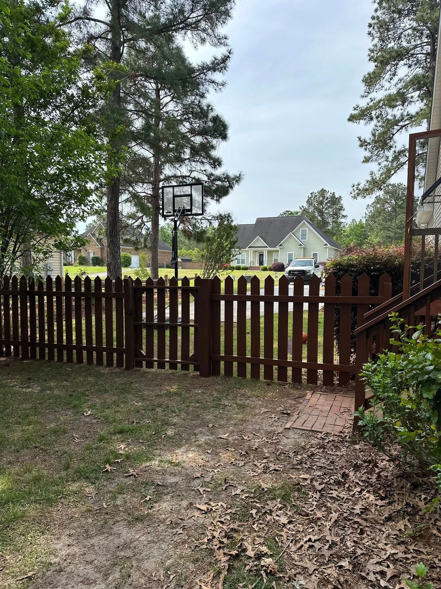 A backyard scene featuring a brown wooden picket fence with a gate, a basketball hoop, and a house in the background.