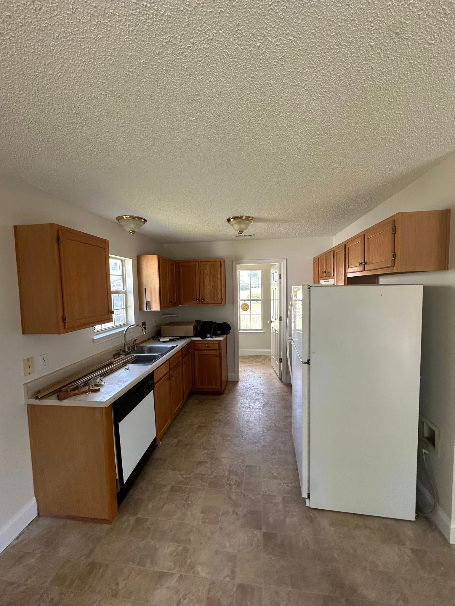 A narrow galley kitchen with light oak cabinets, white appliances, and a textured popcorn ceiling.