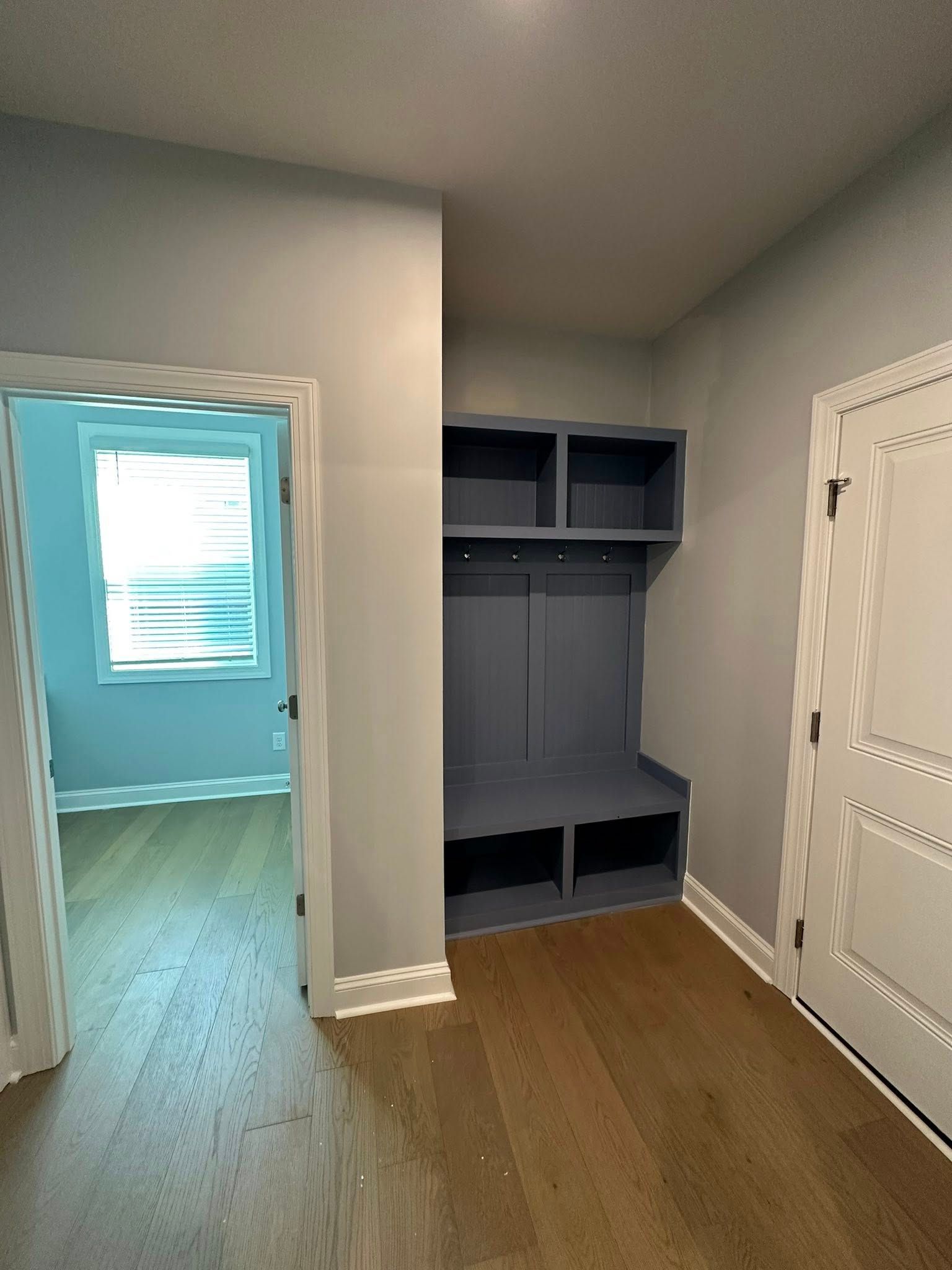 A blue built-in storage bench unit sits in a hallway between a door with trim and an entryway to a light-filled room.