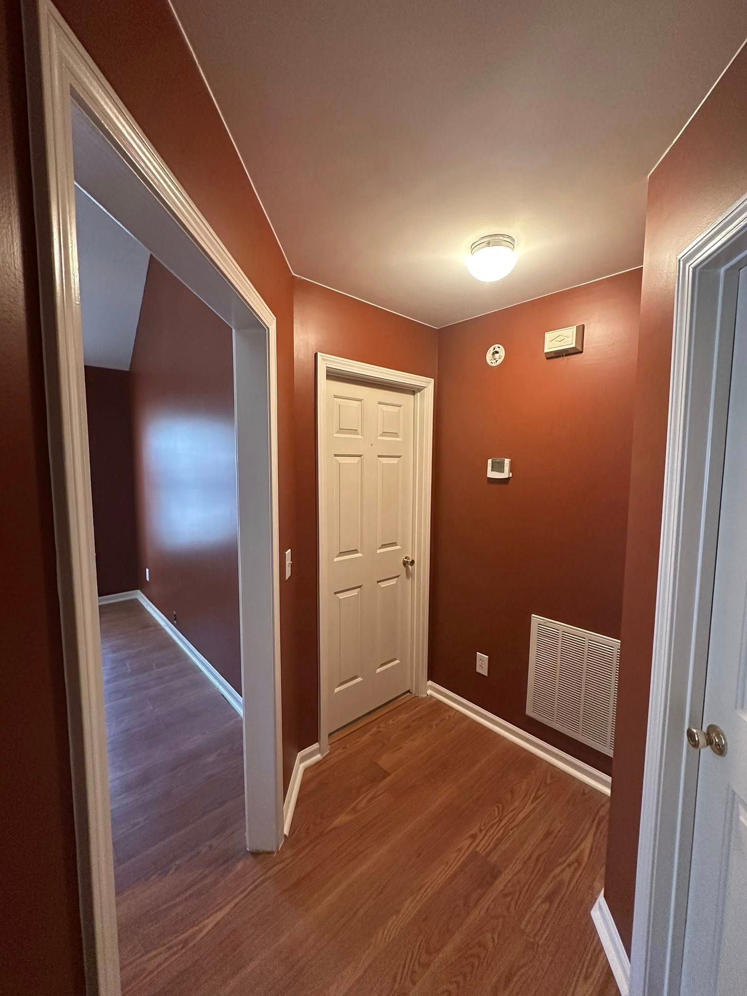 A hallway with dark terracotta walls, hardwood floors, a white door, and a white vent cover on the wall.