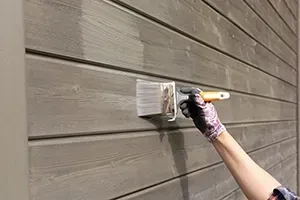 A gloved hand uses a brush to apply gray stain or paint to the horizontal wooden planks of an exterior wall.