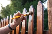 A hand uses a yellow-handled brush to apply wood stain to a wooden picket fence in an outdoor setting.