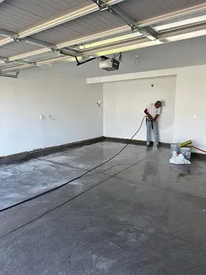 A person uses a pressure washer to clean a large, gray concrete garage floor.