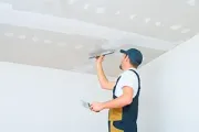 A worker in blue and tan coveralls applies joint compound to a ceiling seam with a putty knife.