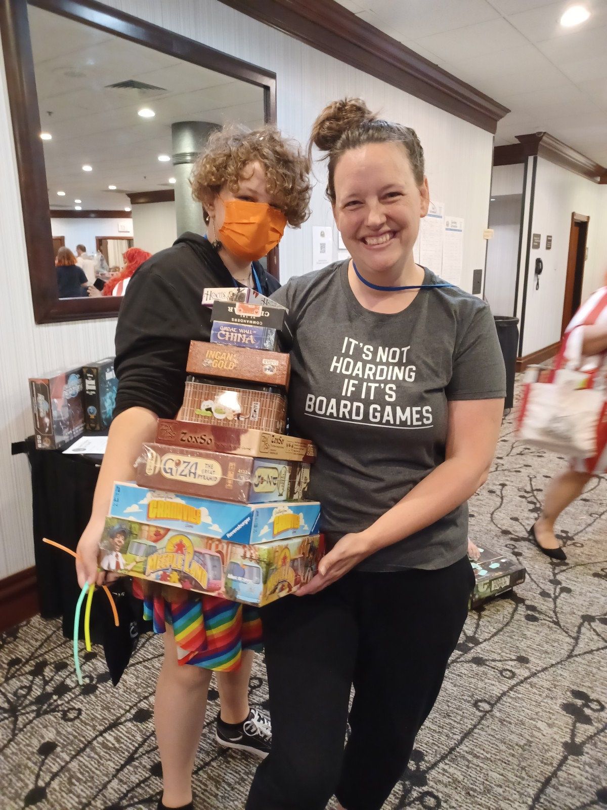 Two women are standing next to each other holding boxes of board games.