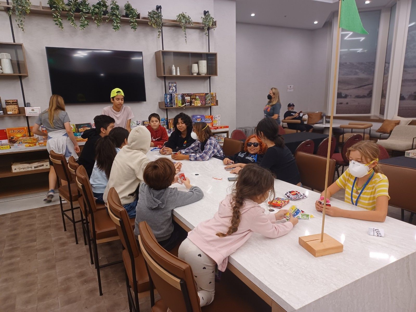 Children at a table building, with adult supervision in a room with plants and a TV.