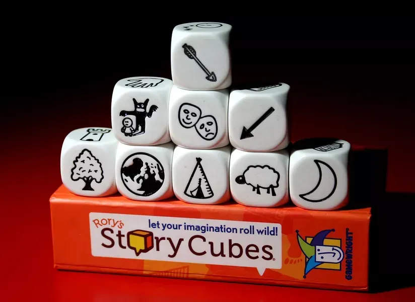 Rory's Story Cubes: white dice with black icons stacked on an orange box, dark background.