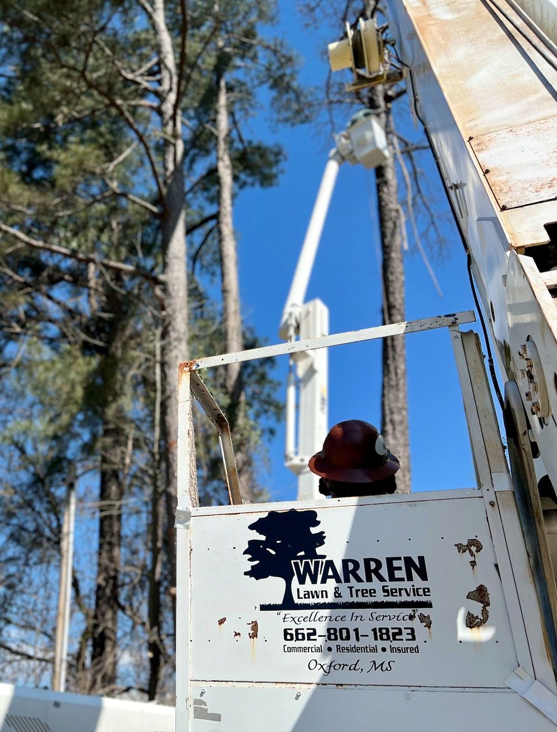 A utility truck with a cherry picker arm extending towards trees on a sunny day. Hard hat sits inside.