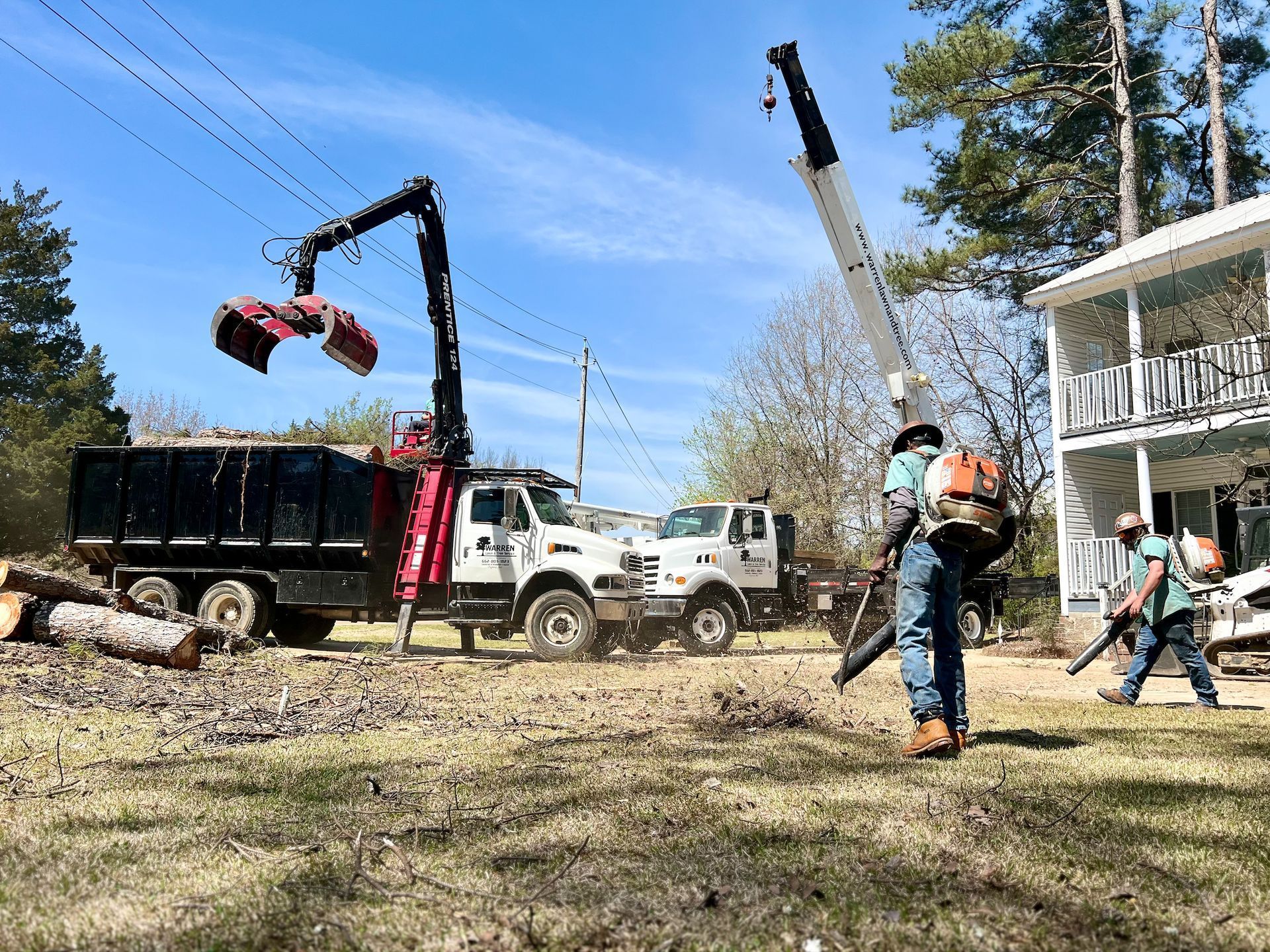 Trucks loading logs, workers using leaf blowers near a house. Bright, sunny day.