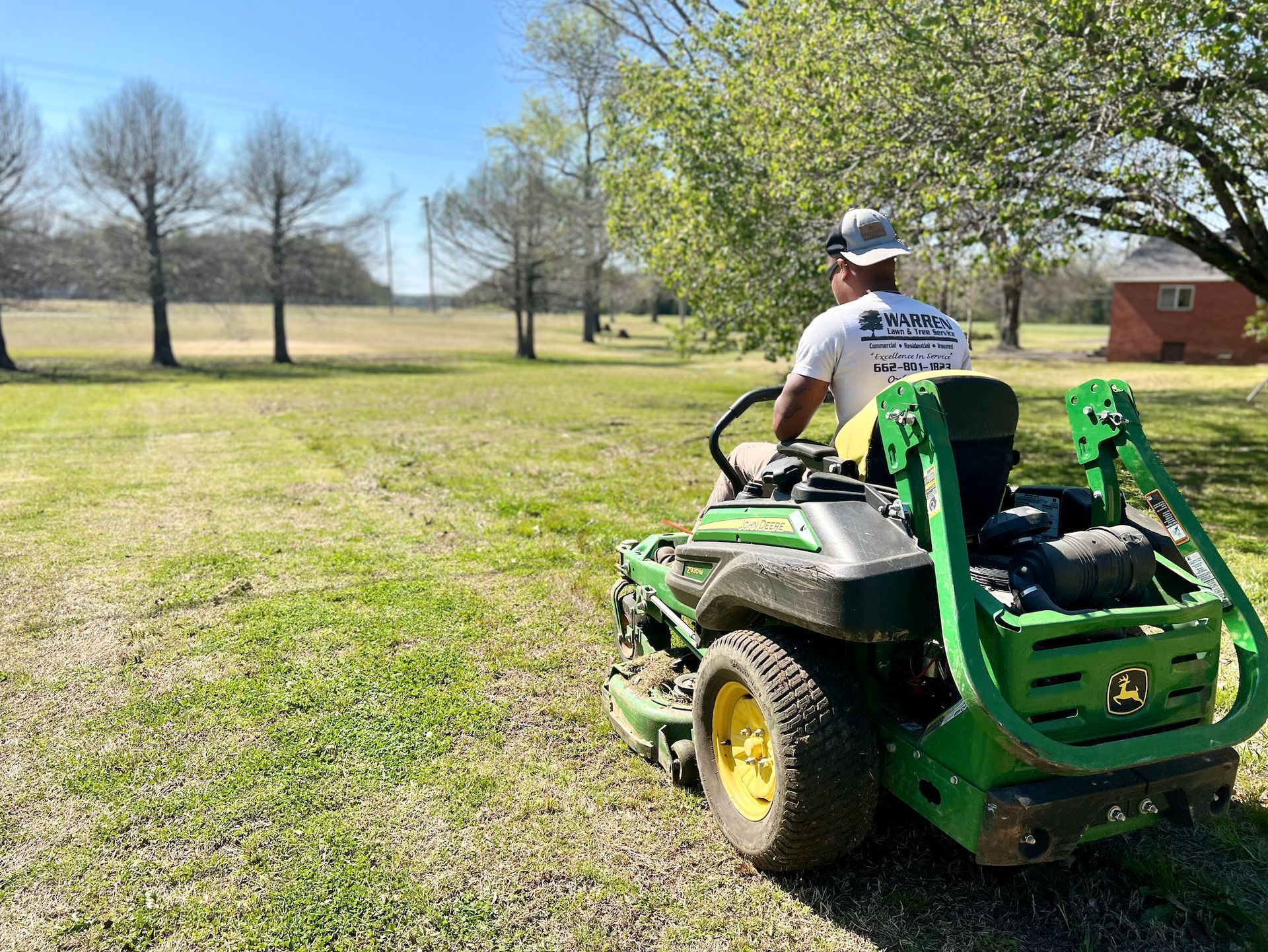 Person on a green John Deere mower cutting grass in a sunny field.