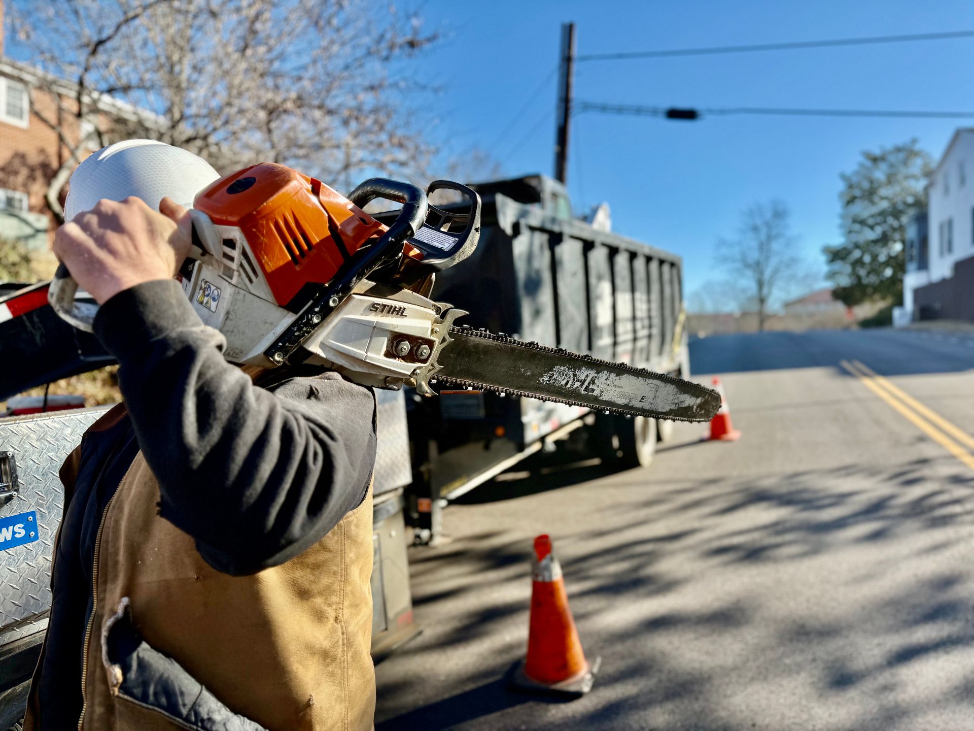 Person holding a chainsaw on shoulder next to truck on street; orange cone in foreground, building in background.