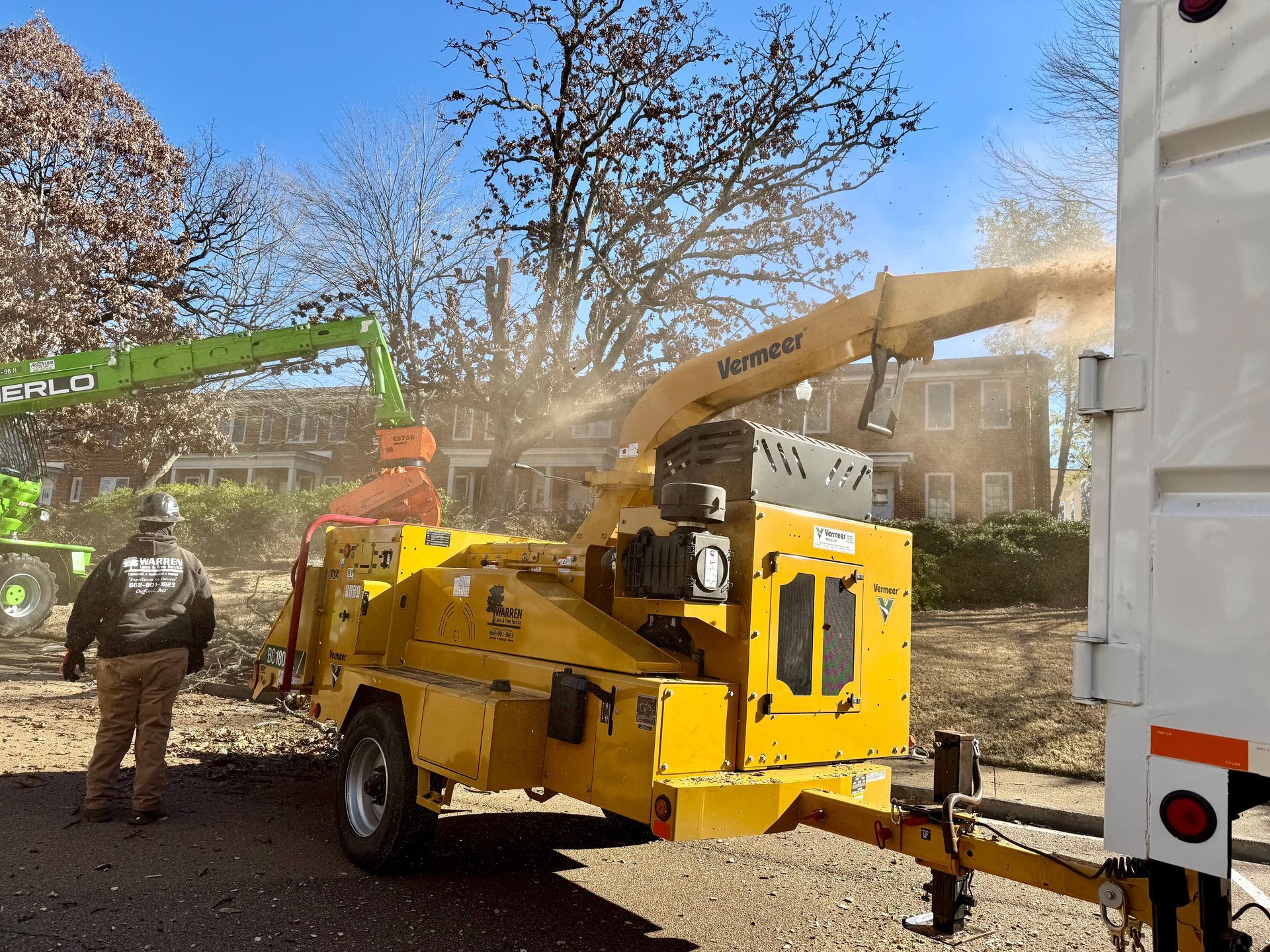 A yellow wood chipper shreds branches; a worker in safety gear stands nearby; green lift and white truck.