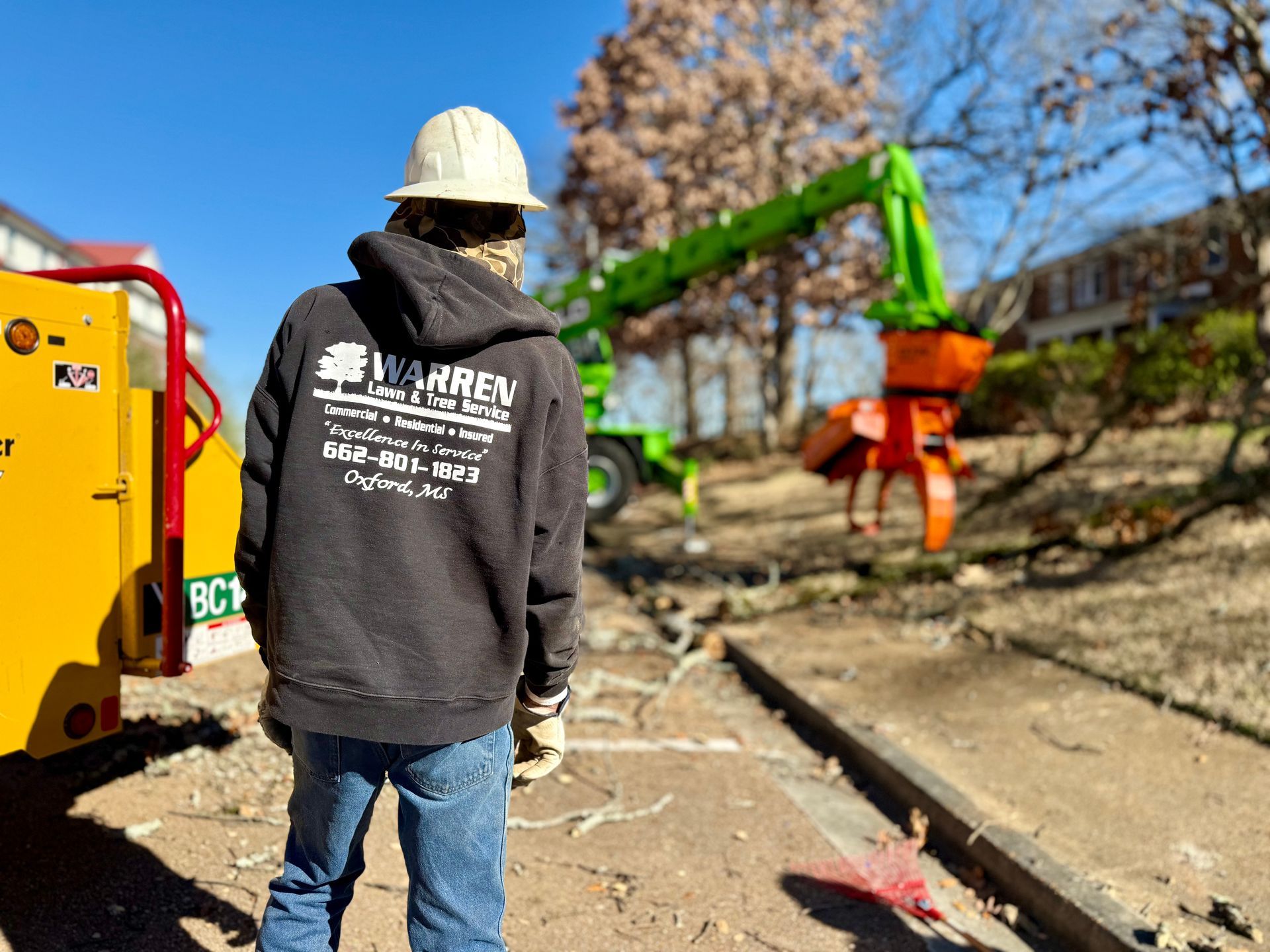 Man in hard hat and black hoodie with logo observing tree removal equipment.