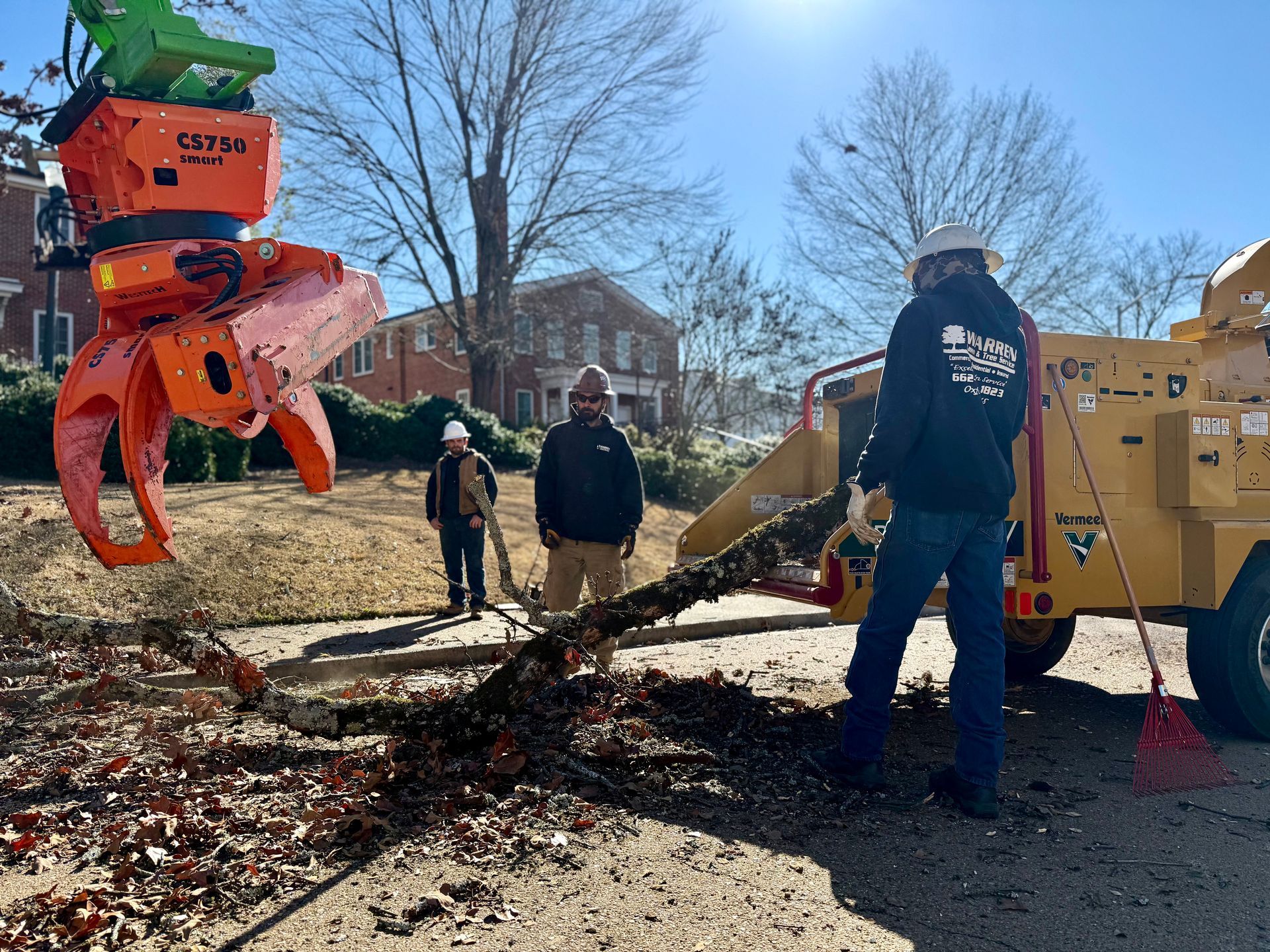Tree removal crew using an orange grapple to feed a yellow wood chipper. Three workers on a sunny day.