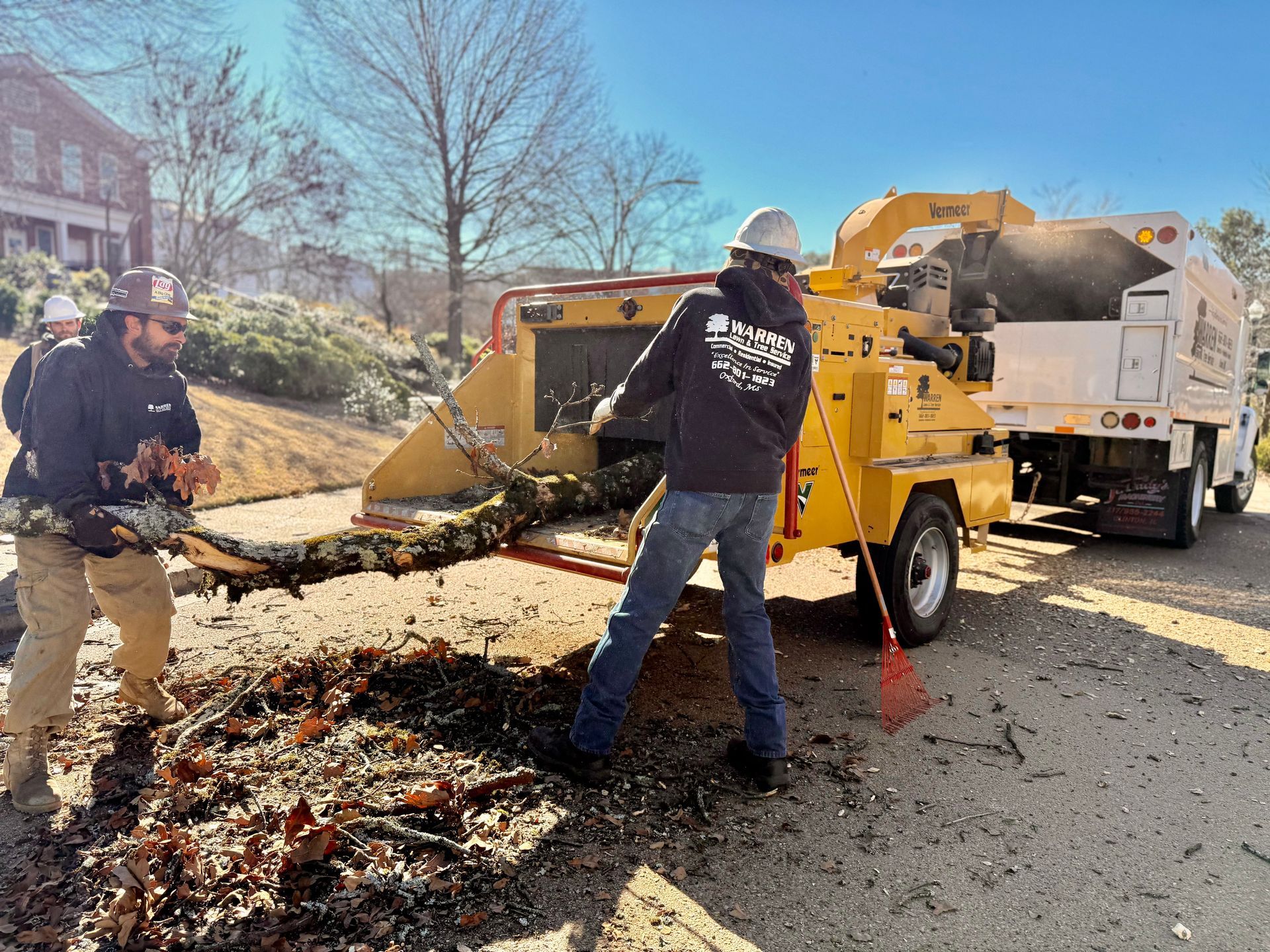 Two workers feeding branches into a yellow wood chipper on a sunny road. A truck is parked nearby.