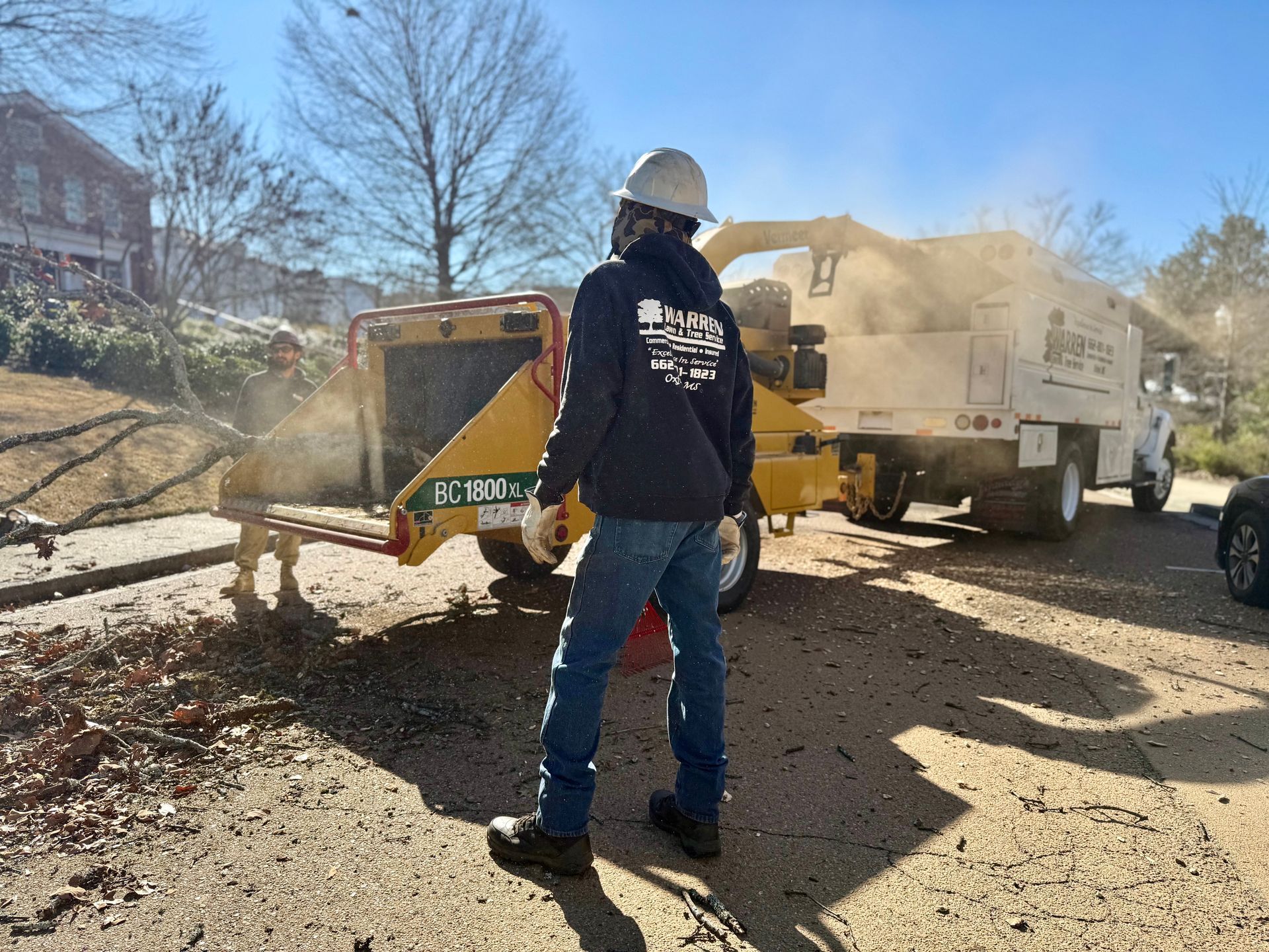Person in hard hat feeds tree branches into a wood chipper; a truck is parked nearby.