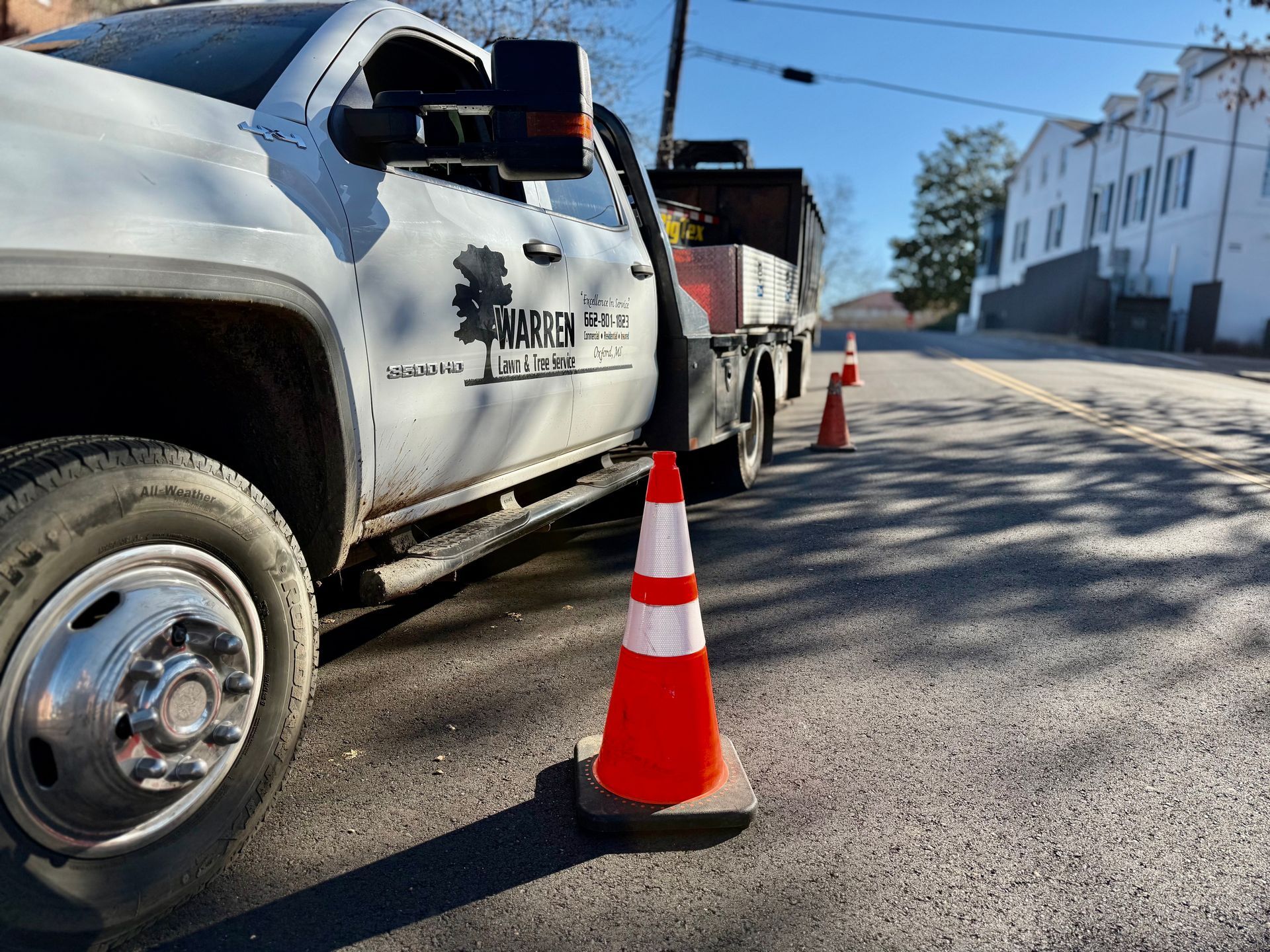 White work truck with orange traffic cones on a street, likely for landscaping work.