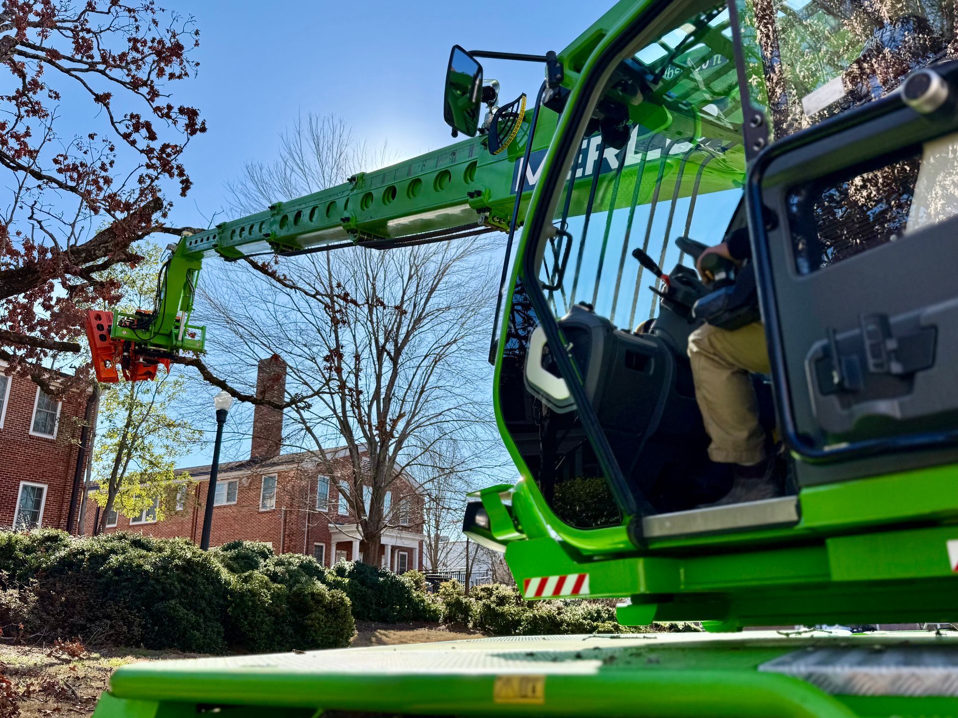 Green tree trimming machine cutting branches; person in the cab. Sunny day, urban setting.