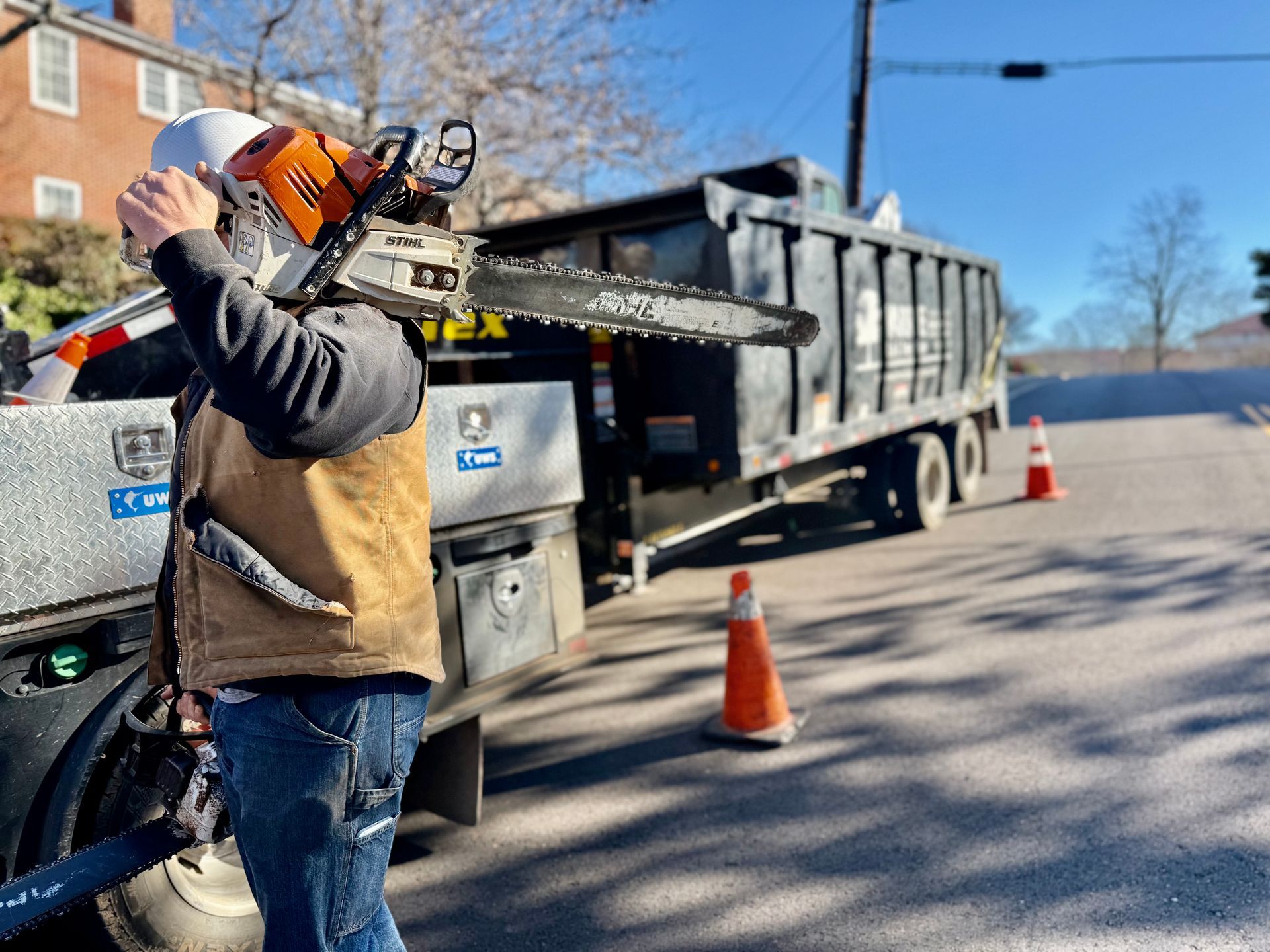 Man with chainsaw near truck on a street, wearing a hard hat and work vest.