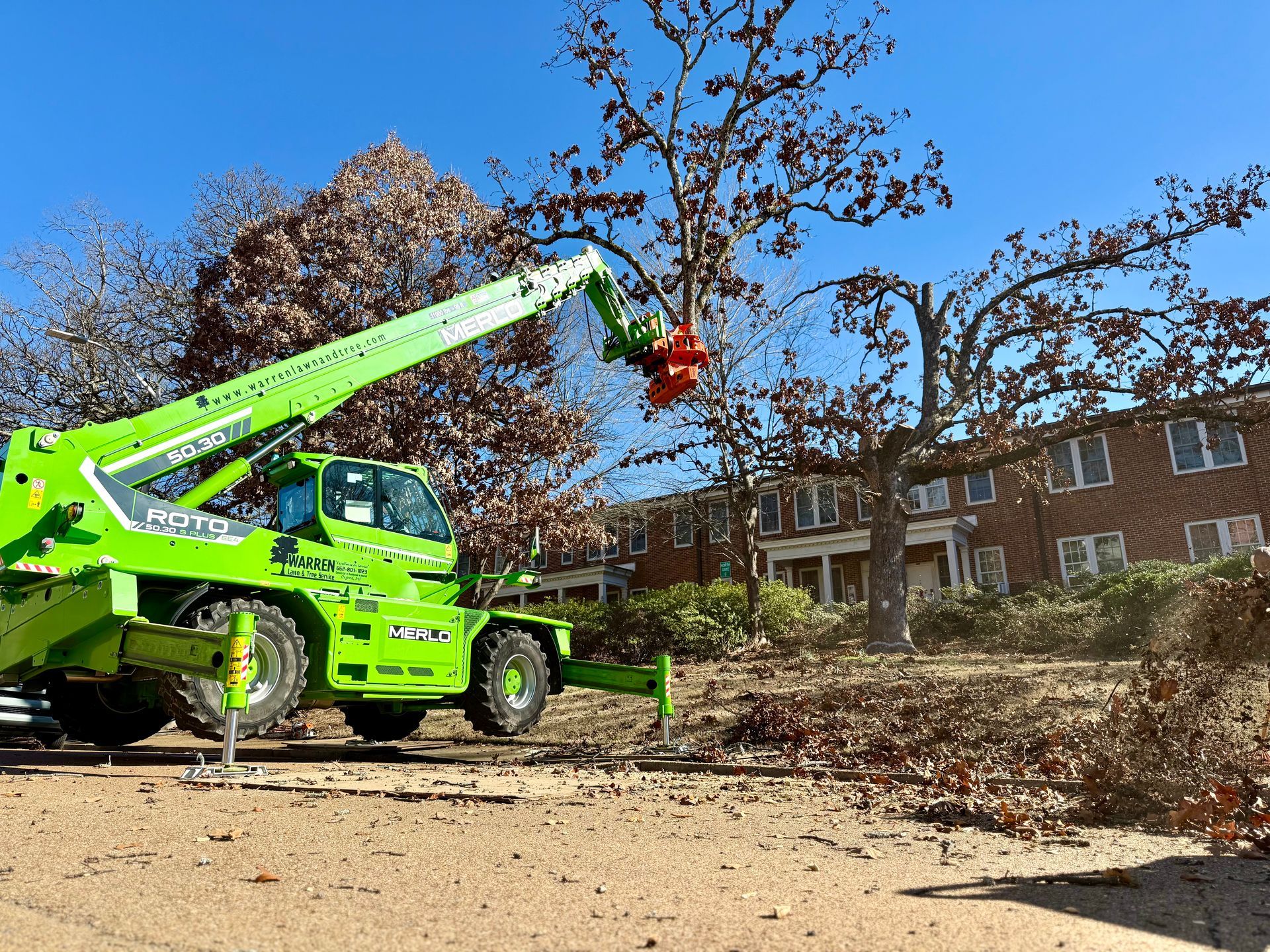 Green tree-trimming machine cutting branches from a tree in front of a building on a sunny day.