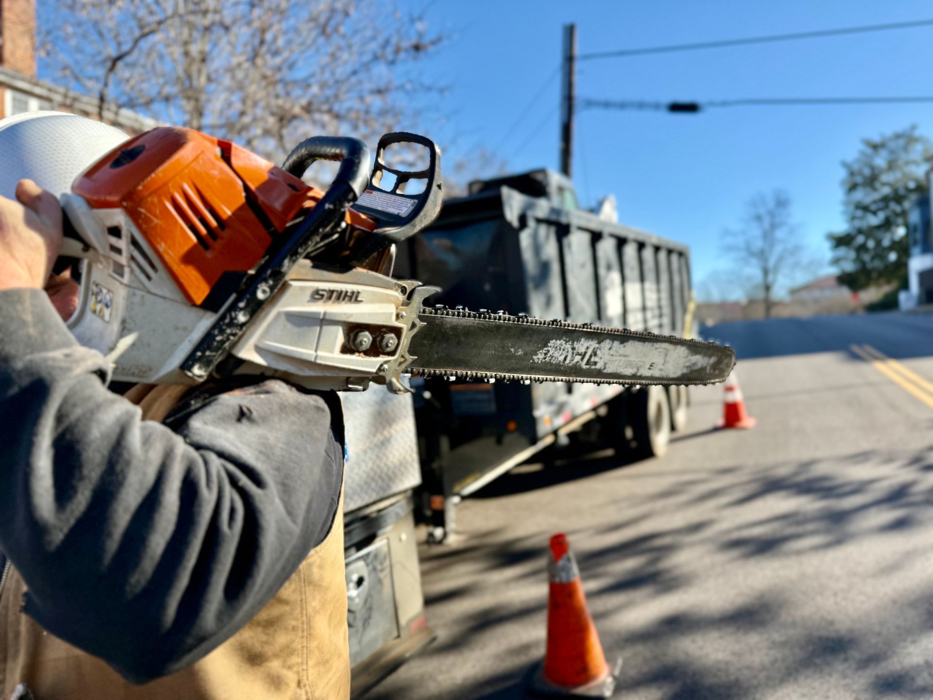 Person holding a Stihl chainsaw near a street with a dump truck and orange traffic cones.