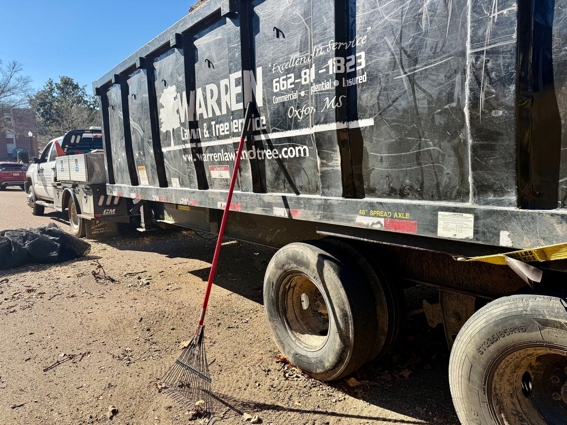 A black dumpster on a trailer with a red broom leaning against it; dirt road setting.