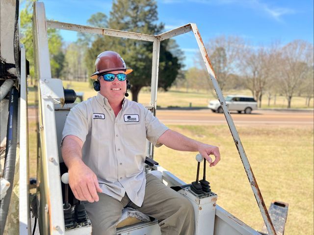 Man in hard hat and sunglasses operates machinery outdoors.