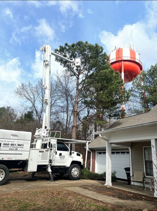 White tree service truck with extended arm near a house, a large tree, and a red and white water tower.