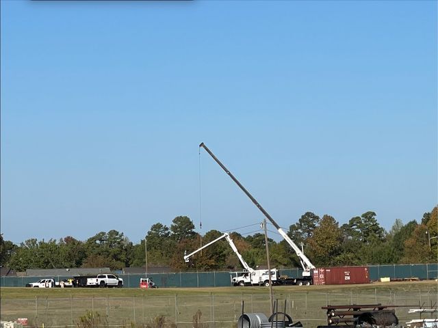 Two cranes lifting objects near a brown container; a blue sky and distant trees are in the background.