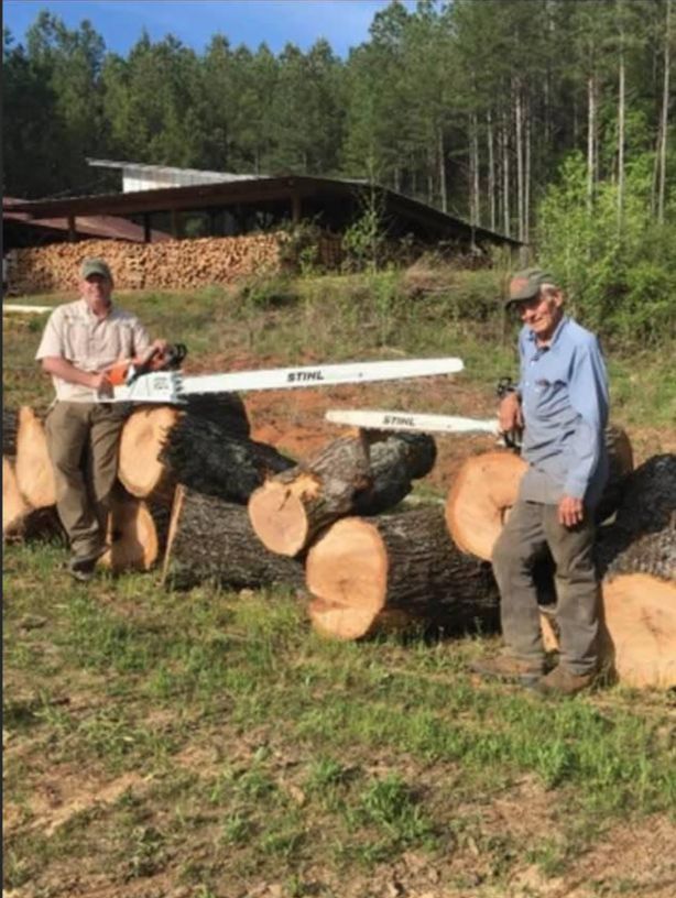 Two men with chainsaws stand near logs, with a woodpile and building in the background.