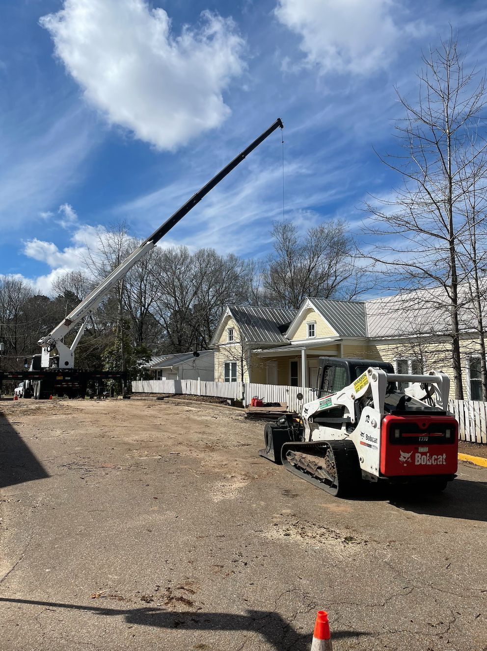 Construction site with crane and Bobcat in front of yellow house under a blue, cloudy sky.
