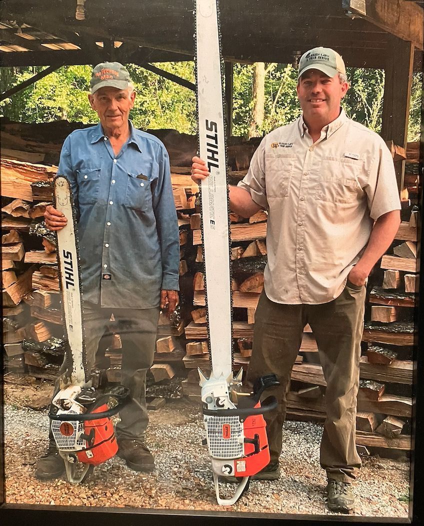 Two men holding Stihl chainsaws in front of stacked firewood. One man wears a blue shirt and the other khaki.