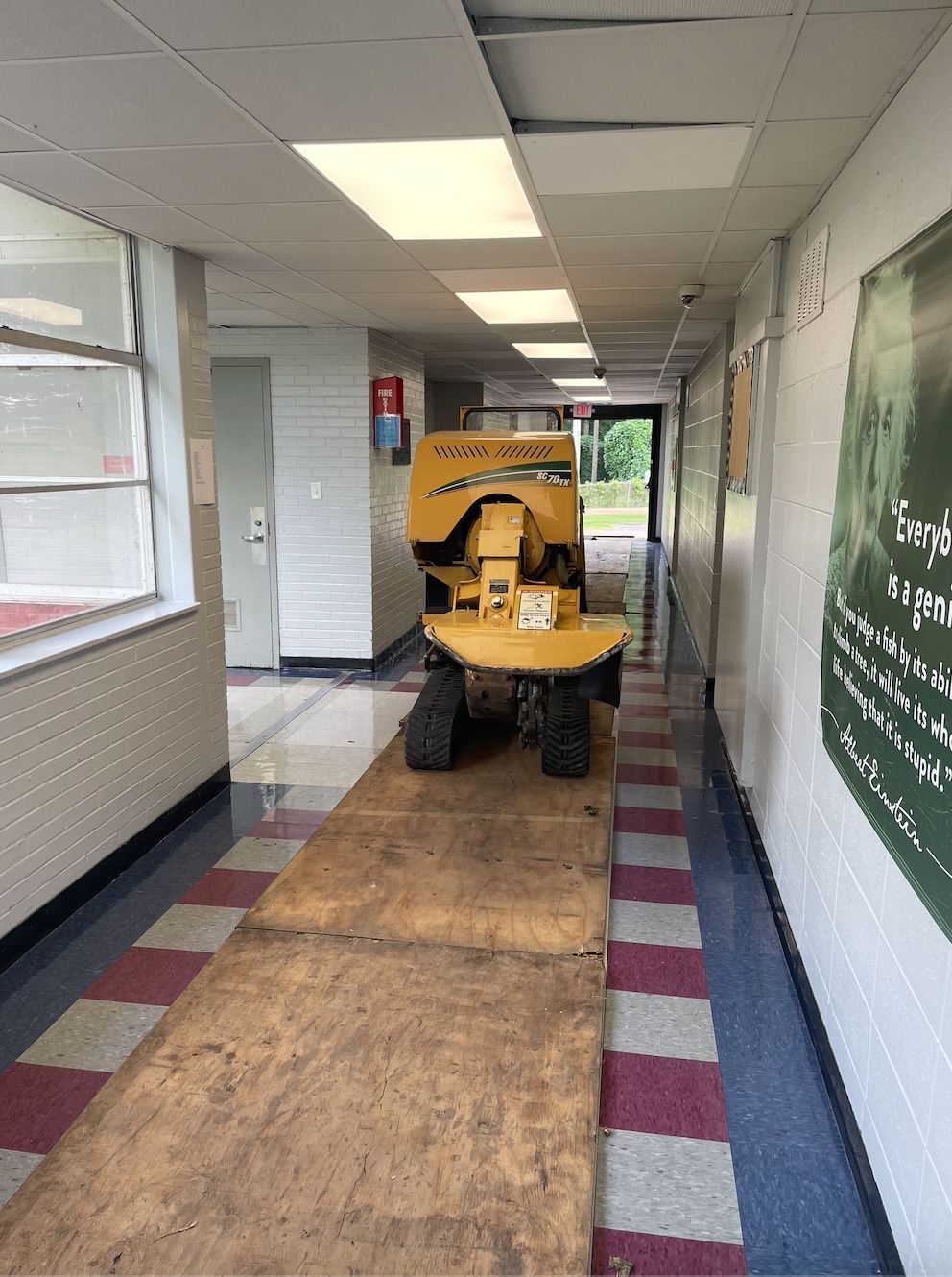 Yellow stump grinder on plywood planks in a school hallway with red and white tile.