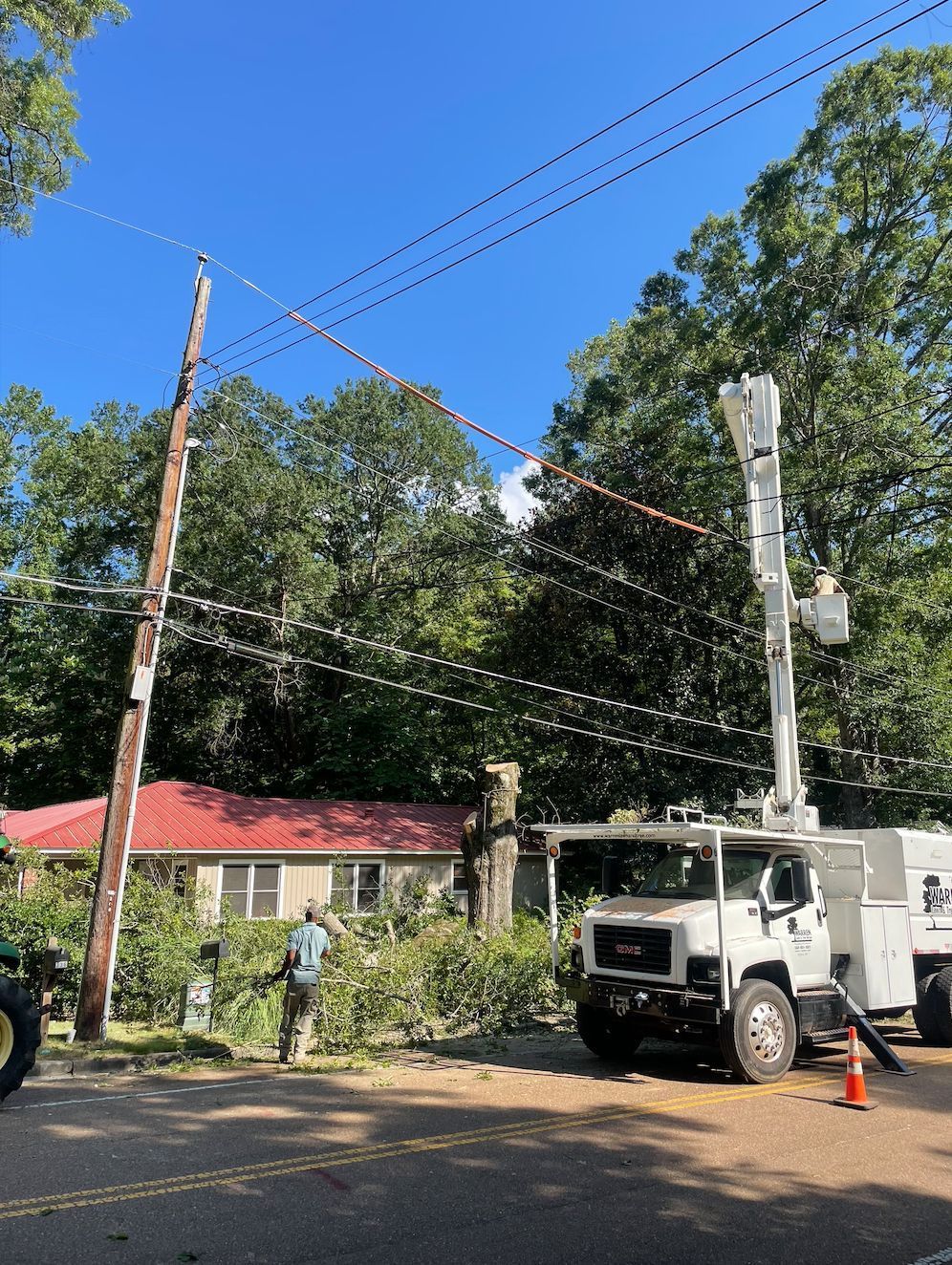 Utility truck with raised boom trims tree near power lines. Person with a chainsaw stands by.