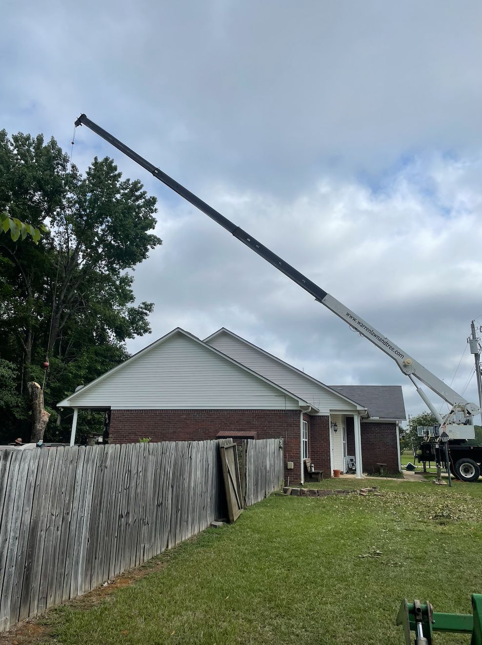 A crane arm reaching over a house, likely for tree trimming. Overcast sky, green grass, and a wooden fence.