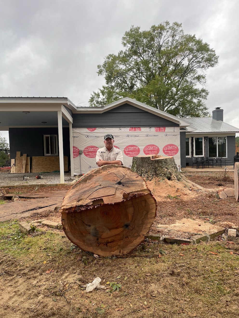 Man stands behind a large tree trunk cut near a house under construction.