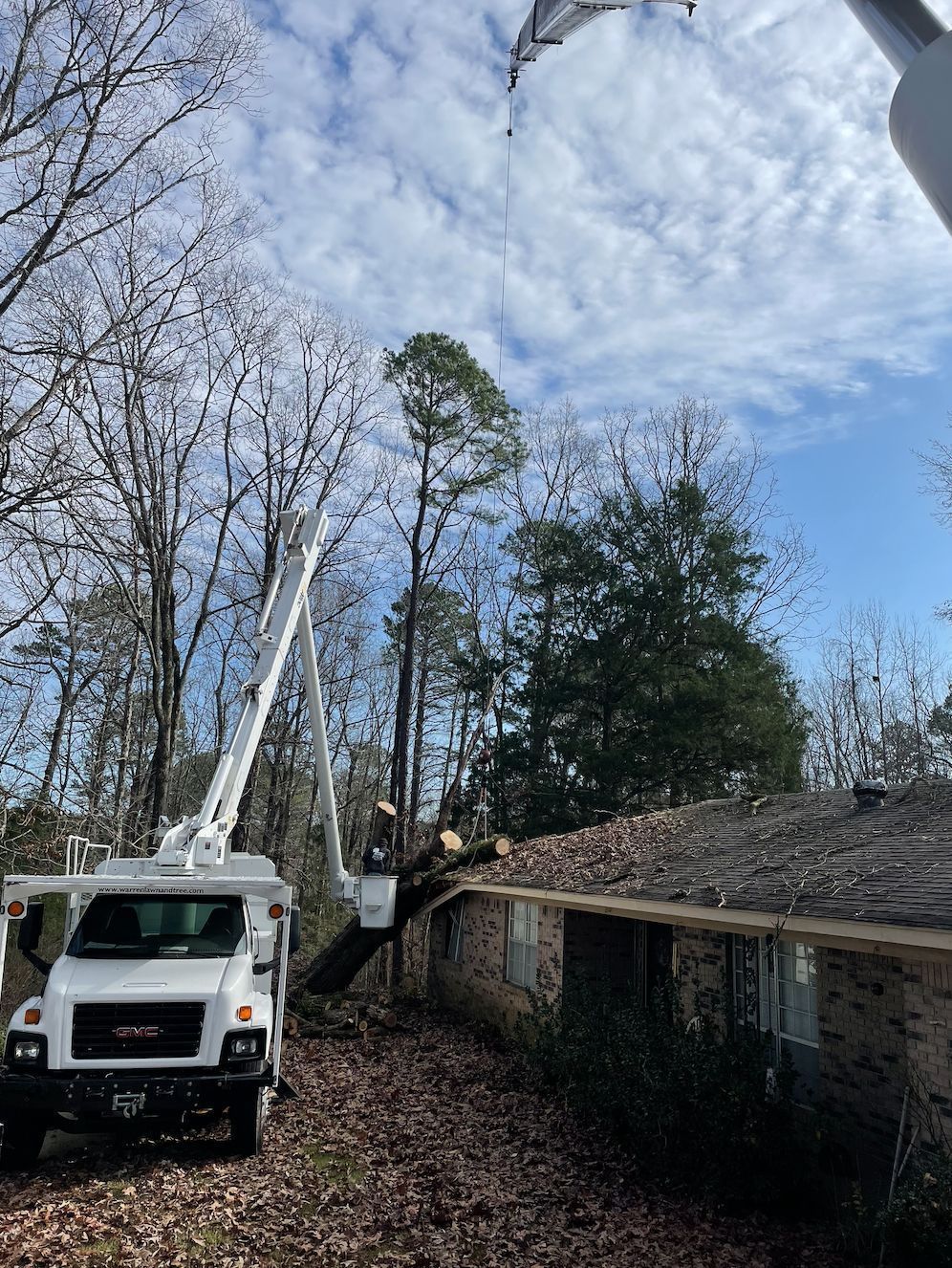Utility truck with extended arm trimming tree branches from a roof. Cloudy blue sky above.