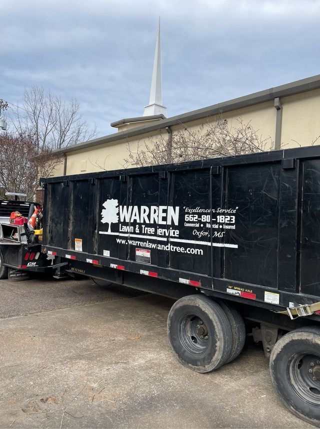 Black dump trailer for Warren Lawn & Tree Service parked near a building with a steeple.
