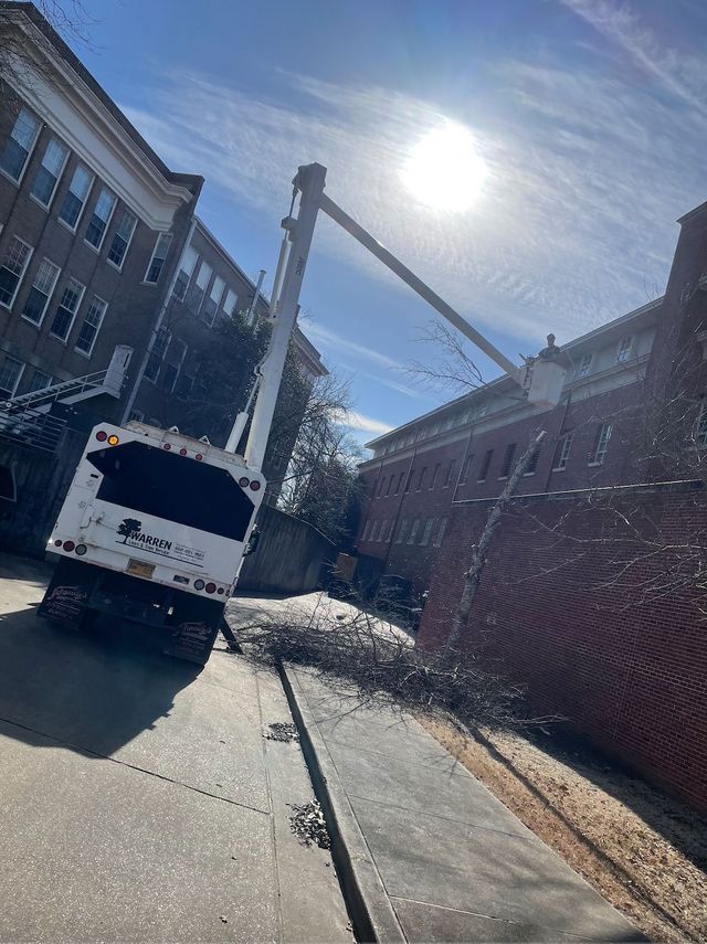 White tree trimming truck with extended arm near a brick building on a sunny day.