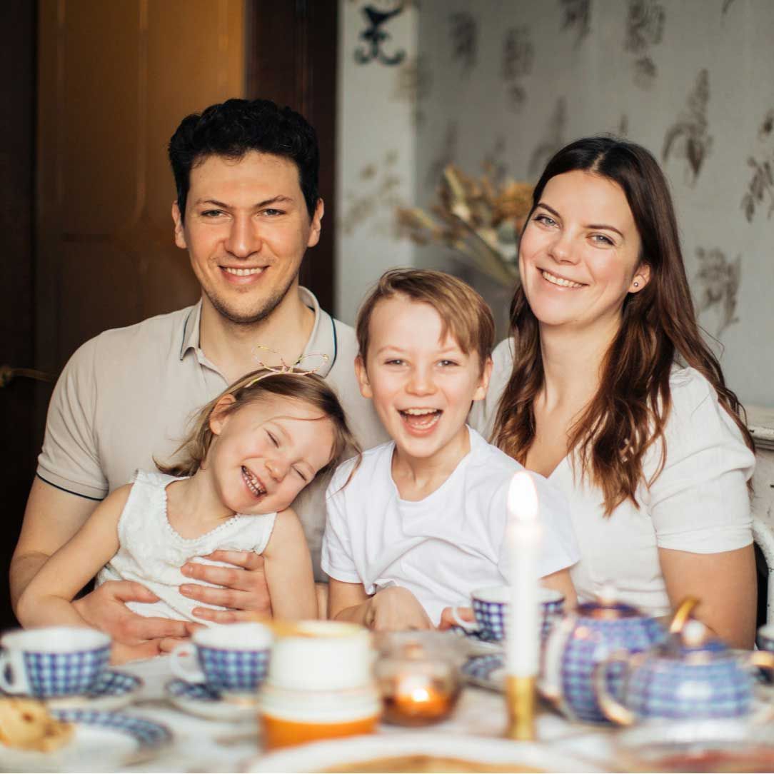 Family smiling at a table set for tea; two children seated with parents.
