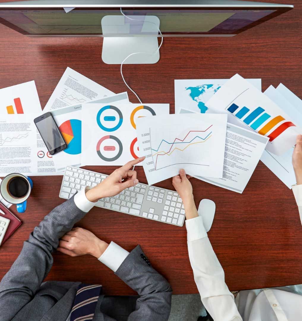 Two people at a desk reviewing charts and graphs, with a computer monitor overhead.
