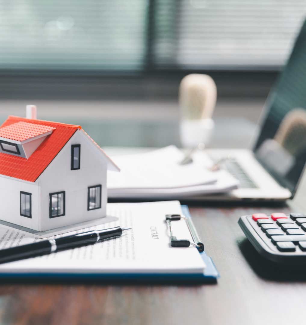 Miniature house model, pen, and paperwork on a desk with a calculator and laptop, suggesting real estate.