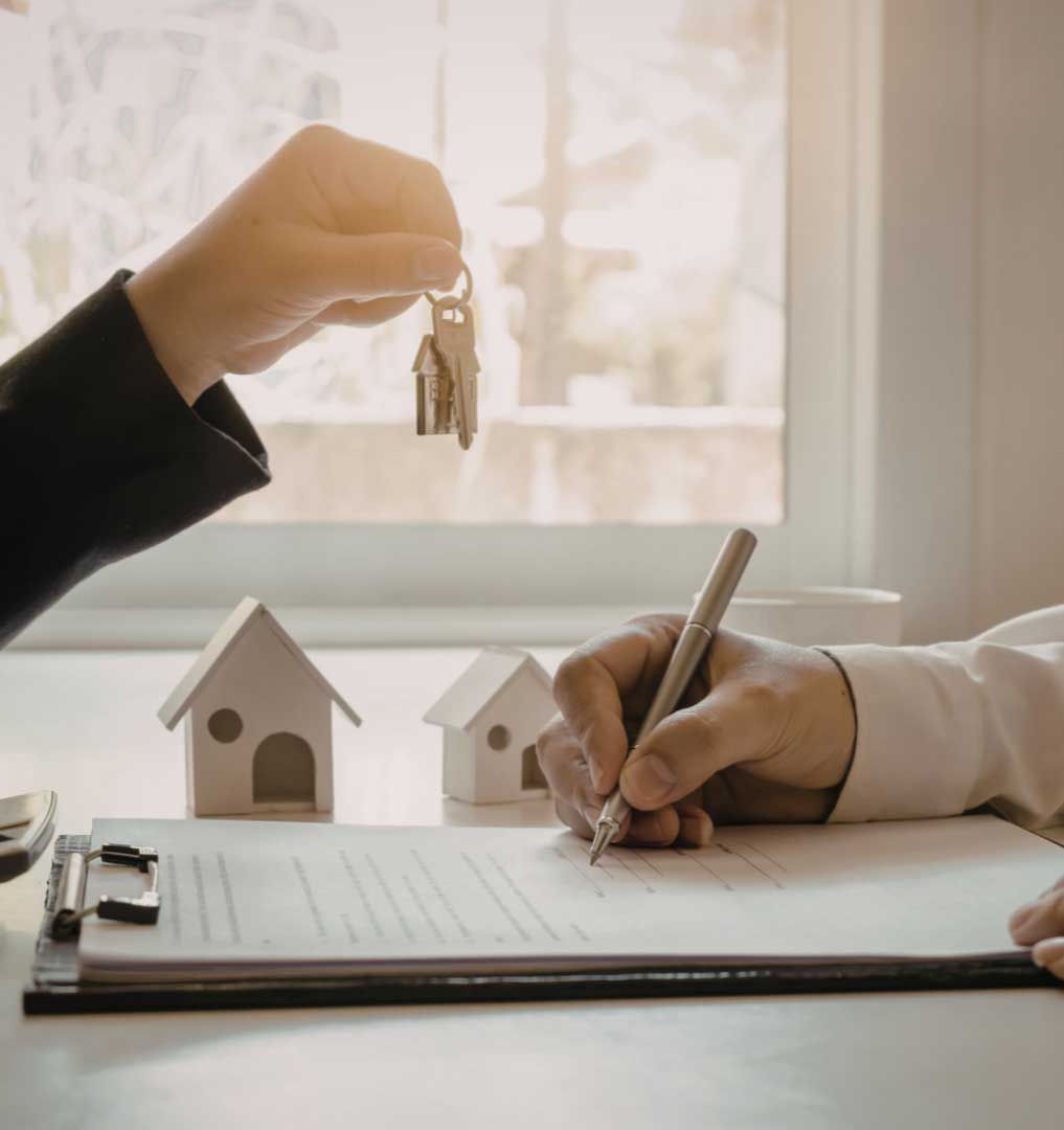 Person handing over keys while another signs a contract on a clipboard, with miniature house models in the foreground.