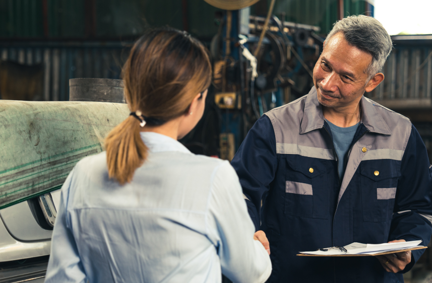 A man and a woman are shaking hands in a factory while the man is holding a clipboard. | Motor Works